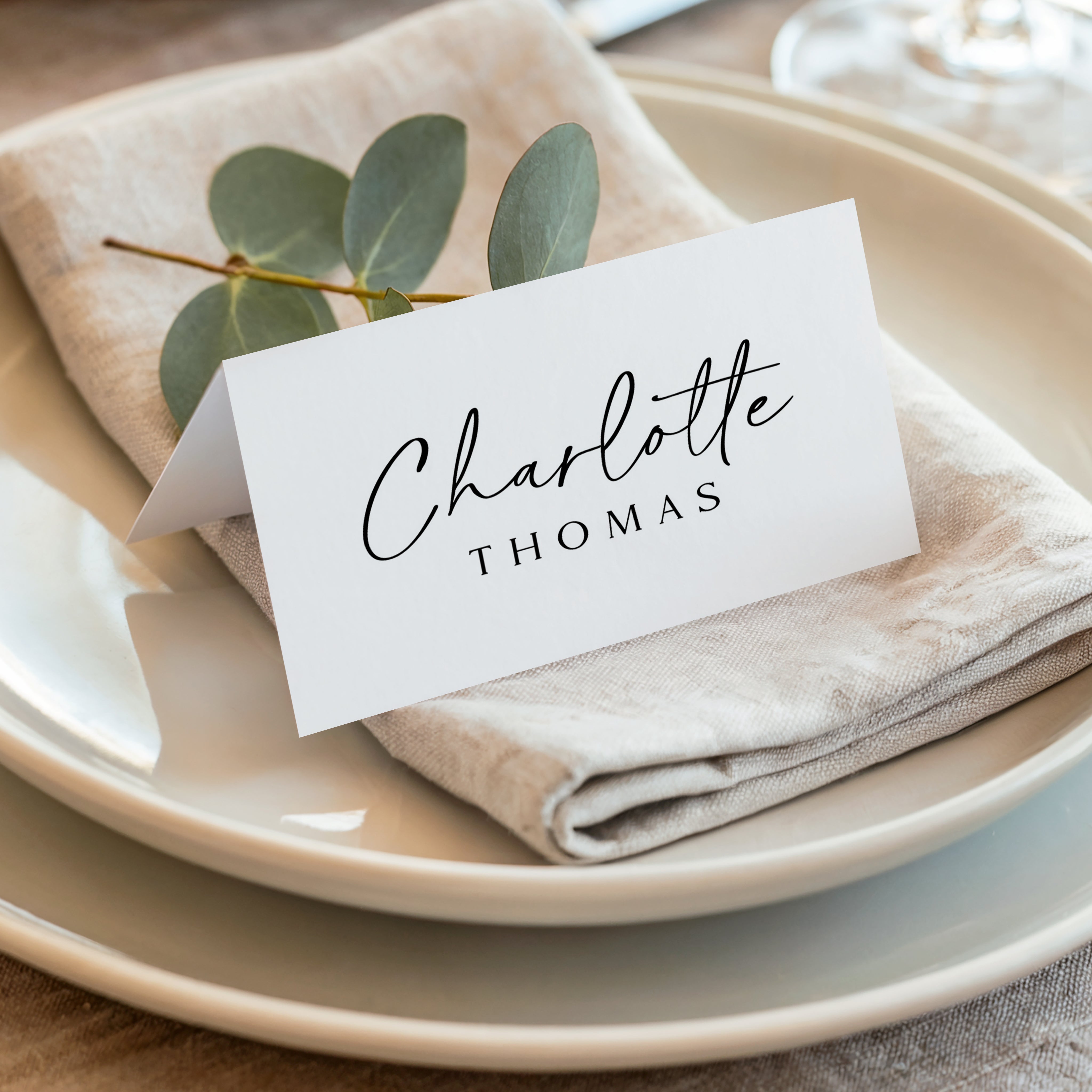White place card with black text that reads 'Charlotte Thomas' on a folded beige napkin with green leaves, set on a white plate.