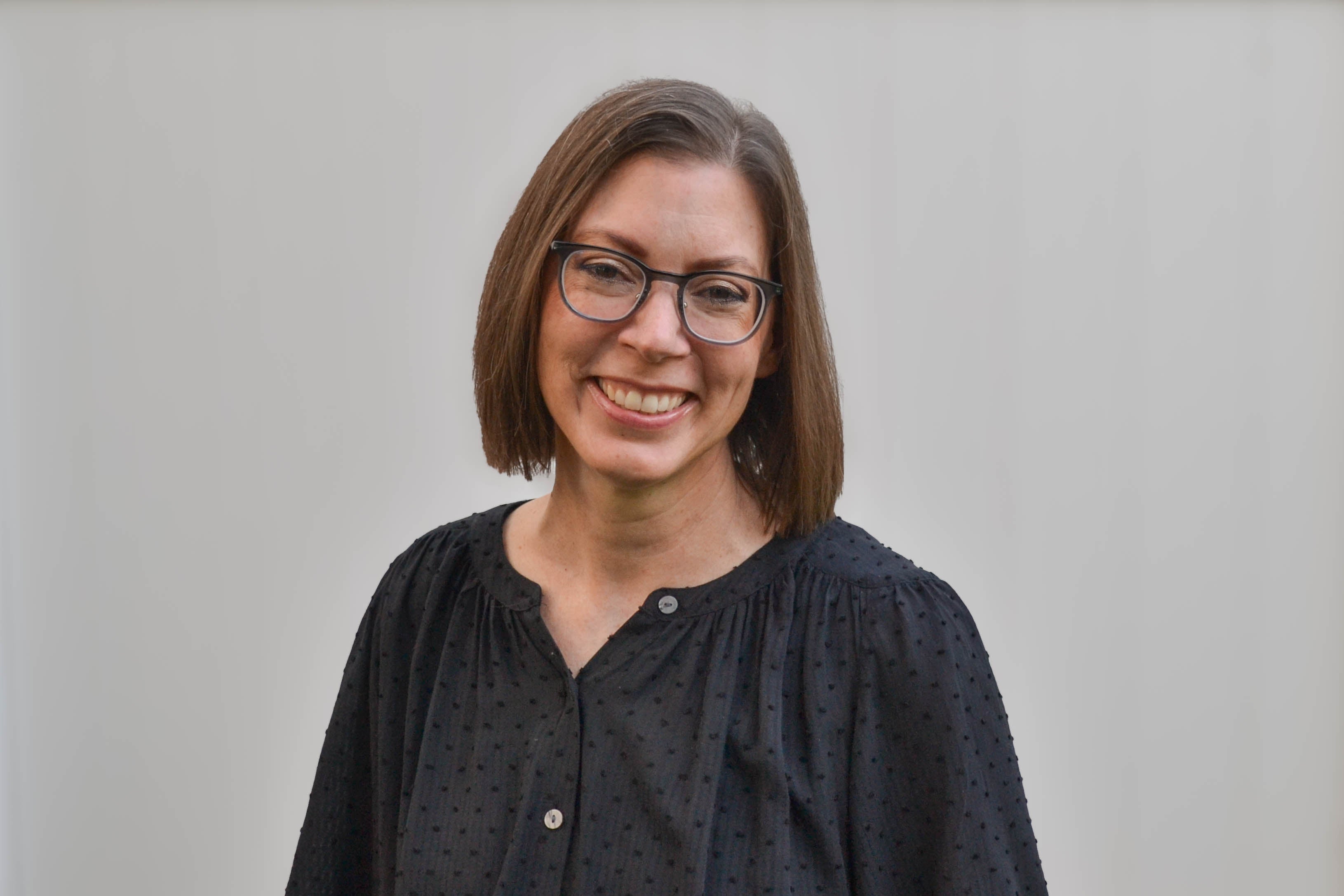 A headshot of Emily, the owner of Olive Invites. She has shoulder length brown hair, wears black frame glasses, and is wearing a black blouse.